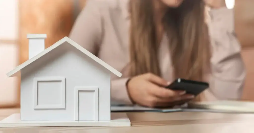 Maqueta de casa sobre mesa con mujer preocupada al fondo revisando su móvil, símbolo de la ocupación de vivienda habitual.